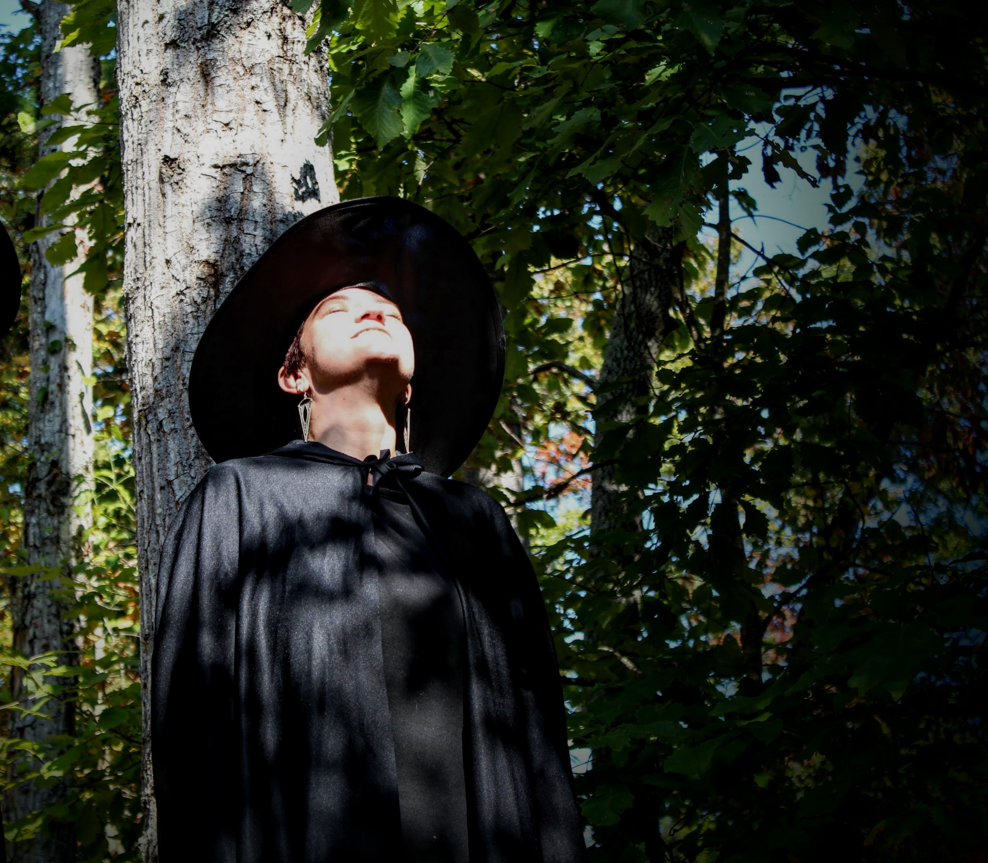 Hannah in a wide black hat and cloak, face turned up to the sun in the woods